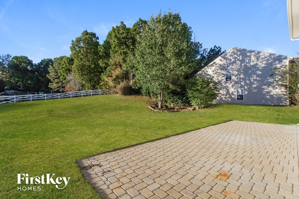 A brick walkway leads to a house with a white fence and green trees in the background.