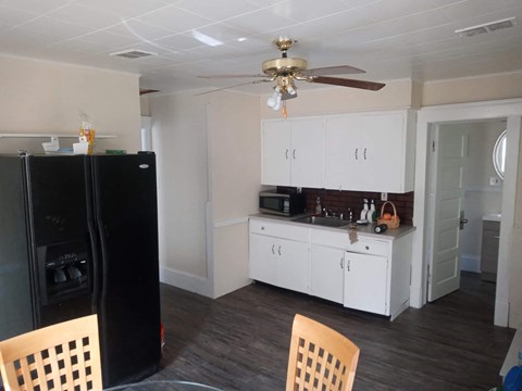 A black refrigerator sits in a kitchen with white cabinets and a ceiling fan.
