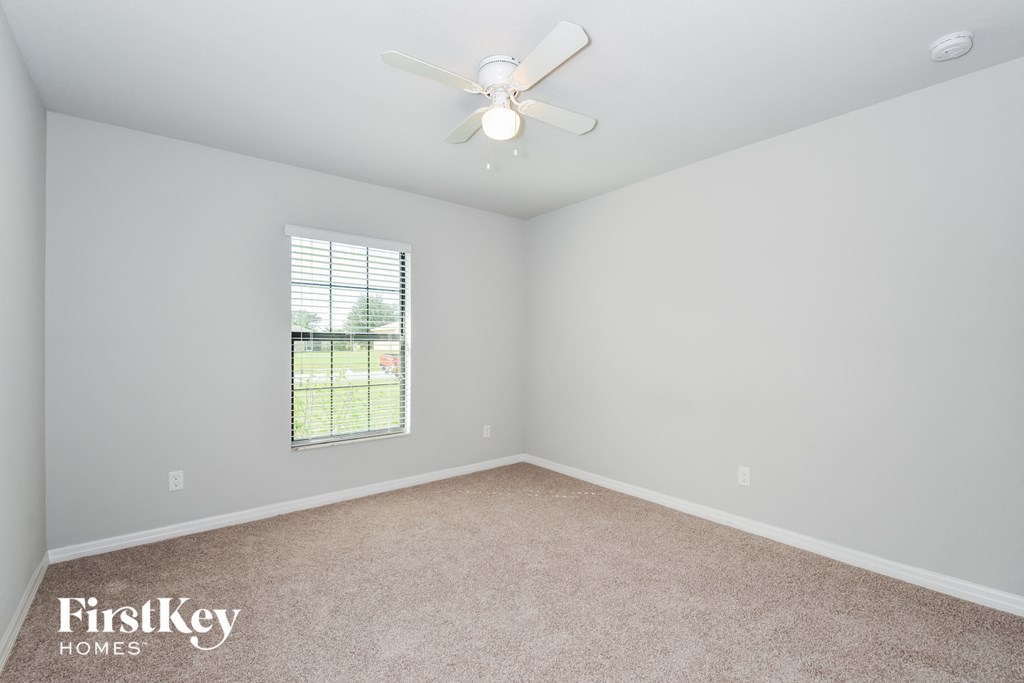 A room with a carpet floor, a ceiling fan, and a window with a view of a fence and trees.
