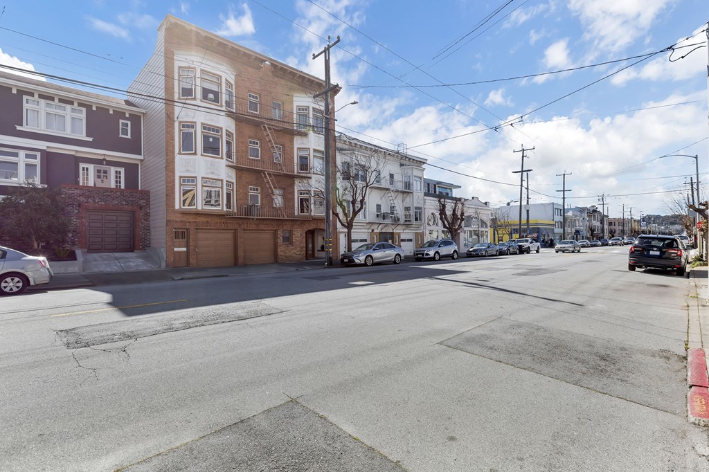 A street view of a residential area with buildings on the left and cars parked on the side of the road.