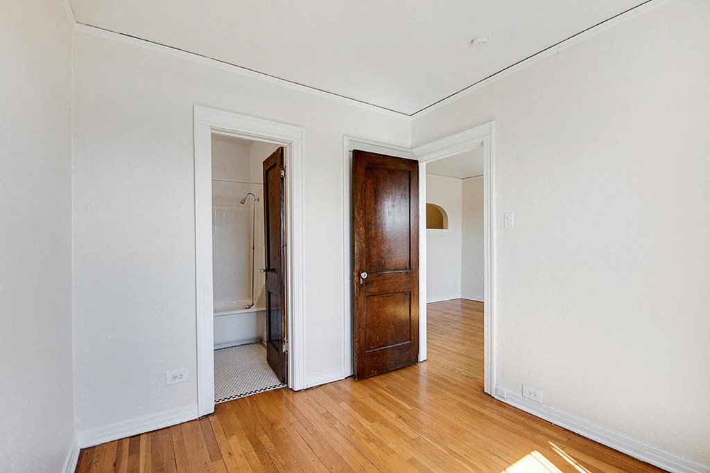 A room with a wooden floor and a white wall with a doorway leading to another room.
