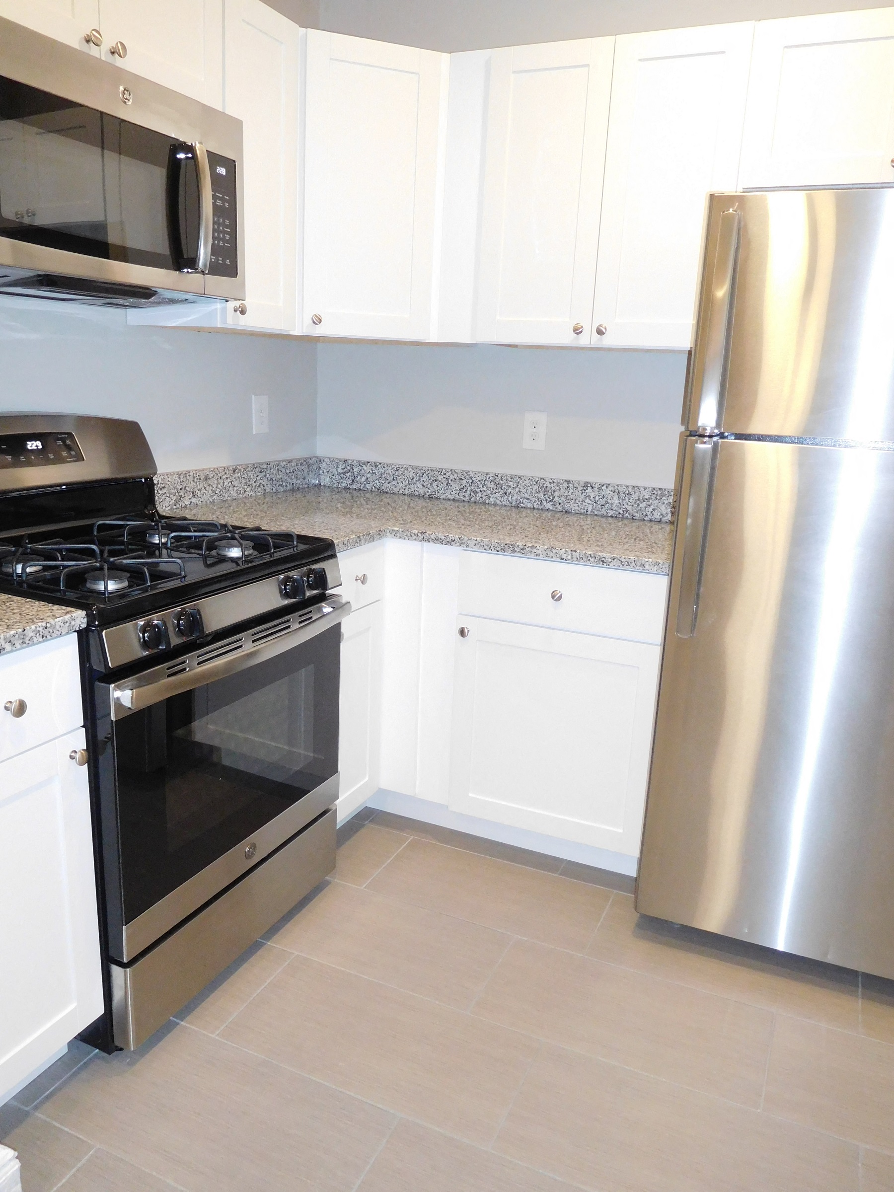 Corner of Kitchen- White Cabinets, Tile Floor, Stainless Refrigerator and Gas Stove with Granite Counters