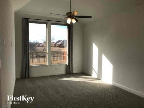A room with a carpet floor, a ceiling fan, and a window with a view of a fence and houses outside.