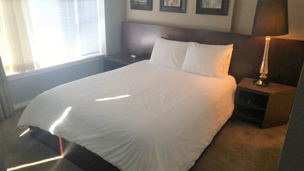 A bed with white sheets and two pillows is in a room with a lamp on a nightstand.