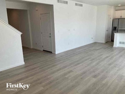 A spacious room with wooden flooring and white walls, with a door on the left and a kitchenette on the right.