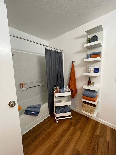 A bathroom with a white door, a shower with a grey curtain, a towel rack with an orange towel, and a shelf with various toiletries.