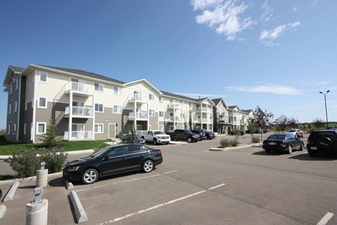 A black car is parked in a parking lot in front of apartment buildings.
