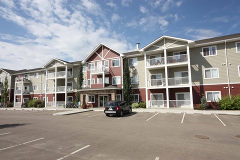 A black car is parked in a parking lot in front of apartment buildings.