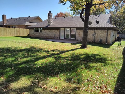 A house with a brown roof and a stone chimney is surrounded by a grassy lawn with fallen leaves.