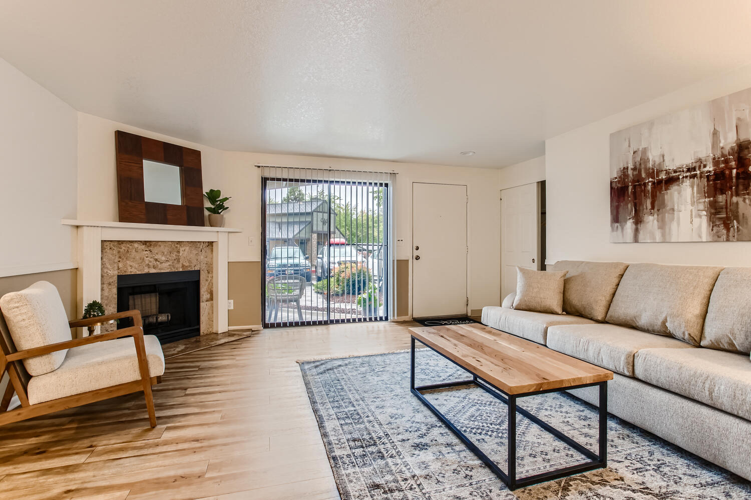 Apartments in Lake Oswego OR - Westlake Meadows - Furnished Living Room with Wood-Style Flooring, Area Rug, Fireplace, Tan Sofa, Armchair, Wooden Coffee Table, and Sliding Glass Door with Patio Access