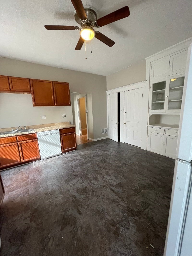 A room with a ceiling fan and a kitchen area with white cabinets.