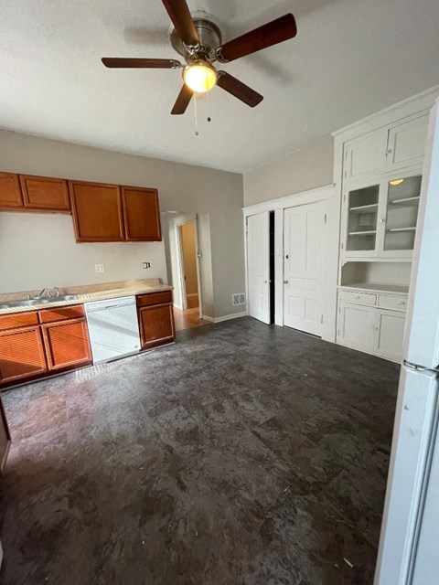 A room with a ceiling fan and a kitchen area with white cabinets.
