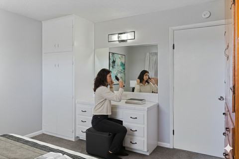 A woman is sitting on a stool in front of a vanity table with a mirror.