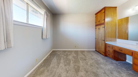 A room with a carpeted floor, a window with curtains, and a wooden cabinet with a white countertop.