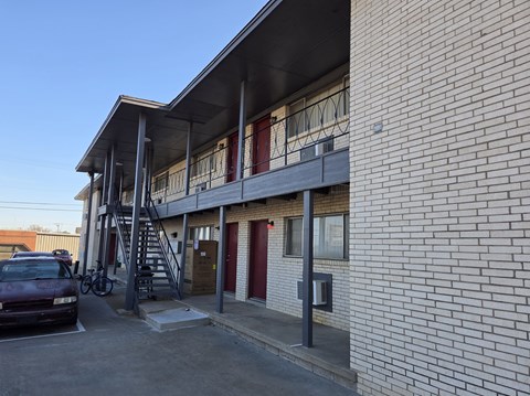 A red car is parked in a parking lot next to a building with a staircase leading to the second floor.