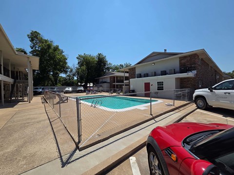 A red car is parked in a parking lot next to a pool.
