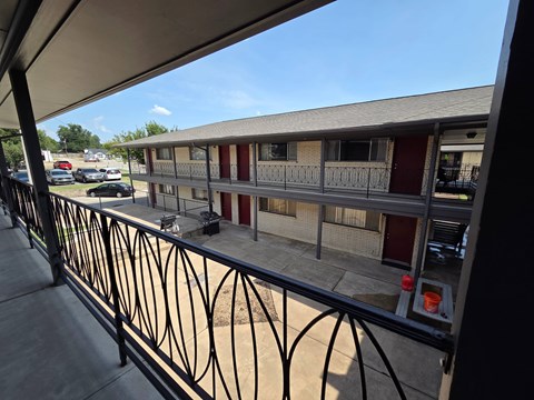 A balcony with a black railing overlooks a parking lot and a building.