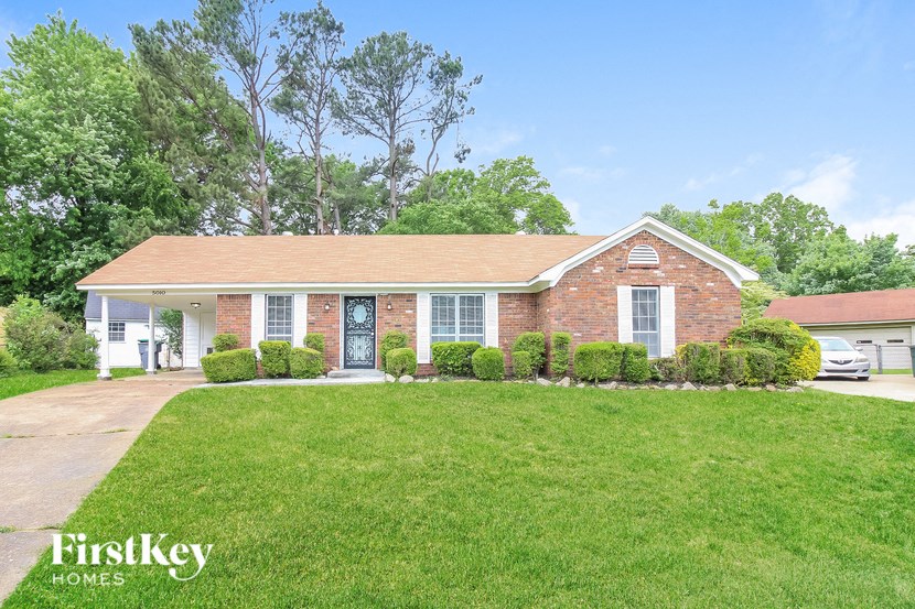 A brick house with a white door and windows is surrounded by greenery.