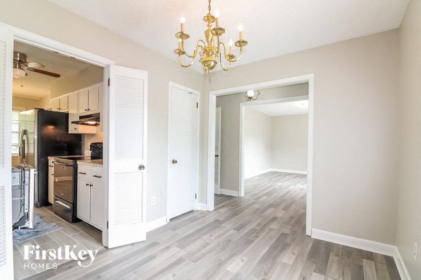 A room with a chandelier and a kitchen area in the background.