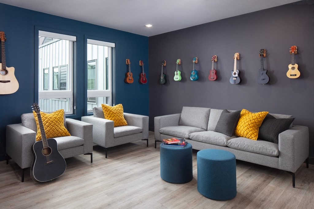 A living room with a grey couch, two chairs, a guitar, and a wall of guitars.
