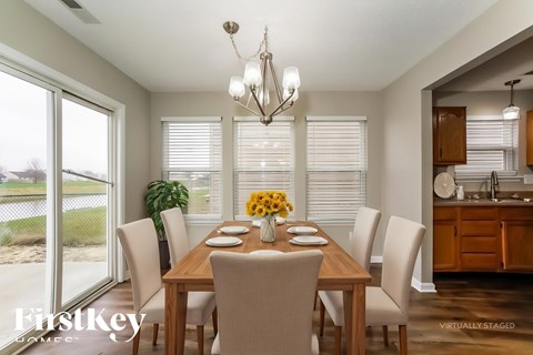 A dining room with a wooden table and chairs, a chandelier, and a kitchen area in the background.