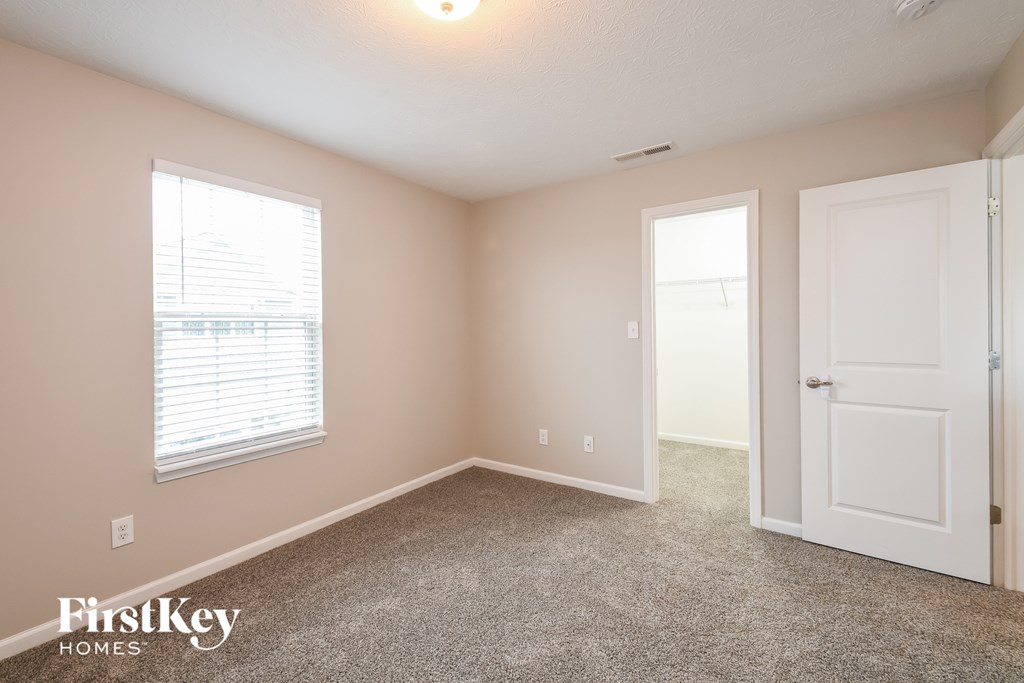 A room with a carpeted floor, a window with blinds, and a door leading to another room.