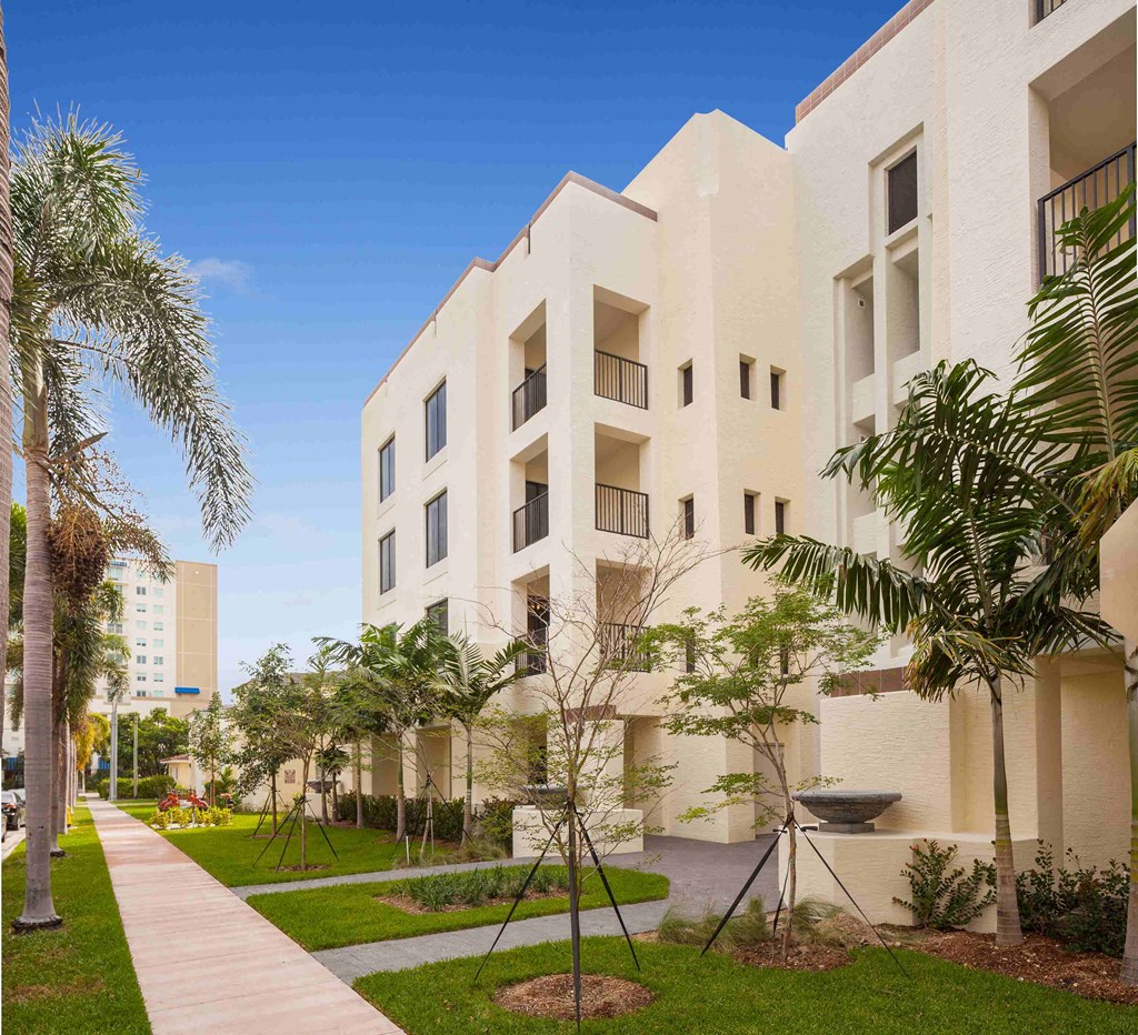 A modern building with a white exterior and balconies is surrounded by greenery and a walkway.