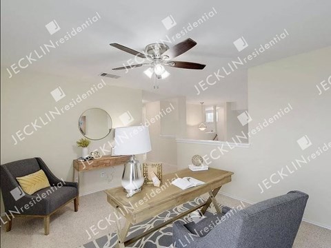 A living room with a ceiling fan, a chair, a table with a lamp and a vase on it, and a mirror on the wall.