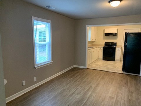 A kitchen area with a black fridge, a window with blinds, and a wooden floor.