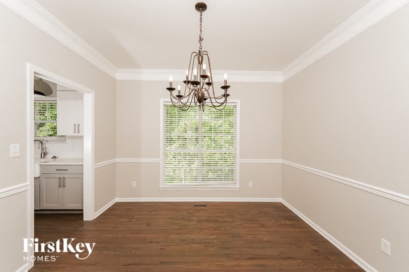 A chandelier hangs in a well-lit room with wooden floors and a window with blinds.