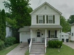 A white house with a grey roof and a black trash bin.