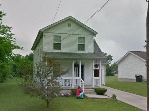A man sits on the front porch of a two-story house.