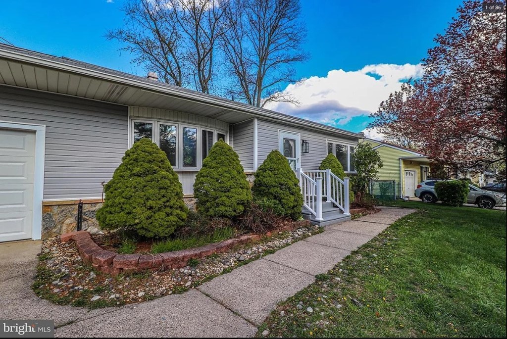 A house with a grey exterior and a white door is surrounded by a small garden with a red brick border.
