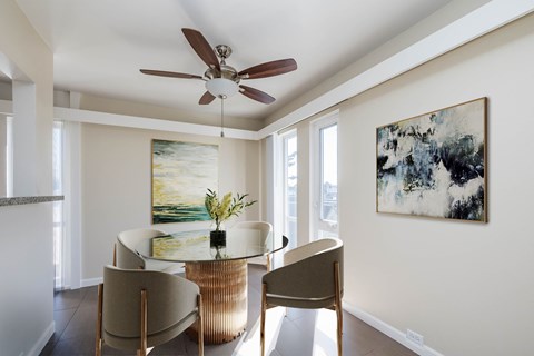 A room with a ceiling fan, a table with chairs, and two paintings on the wall.
