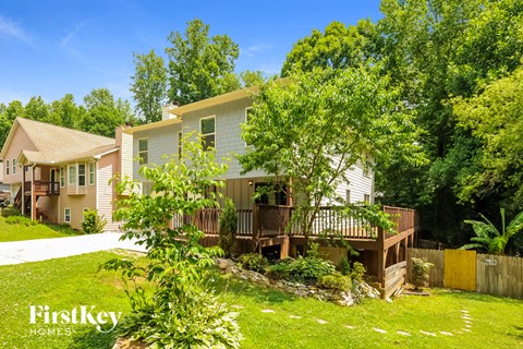 A house with a white exterior and a brown deck is surrounded by greenery.