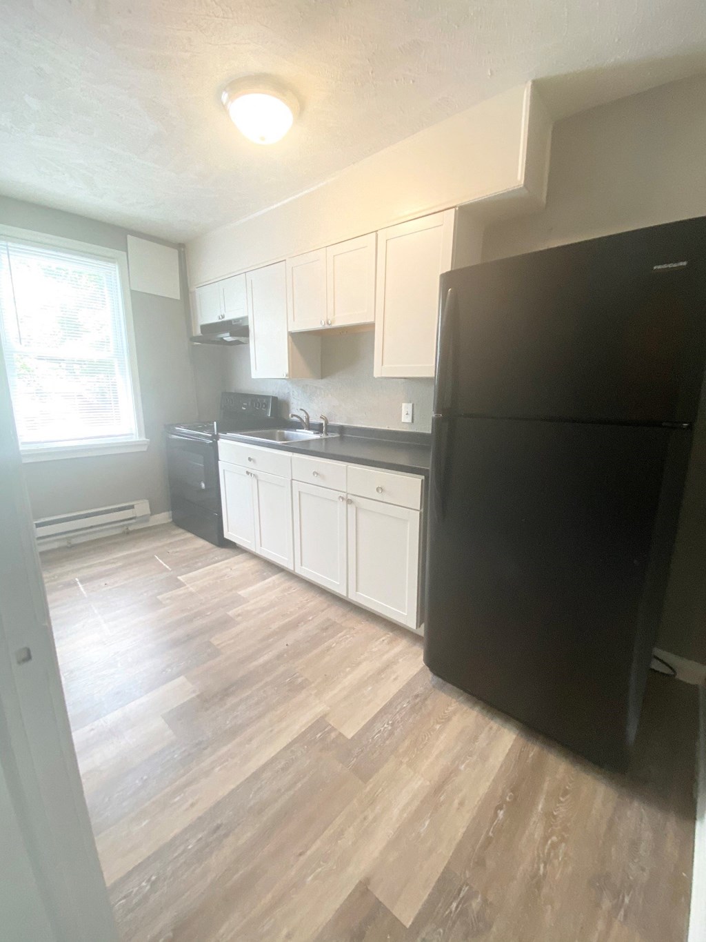 A black refrigerator sits in a kitchen with white cabinets and a wooden floor.