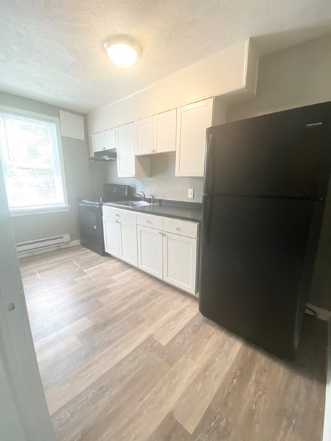 A black refrigerator sits in a kitchen with white cabinets and a wooden floor.