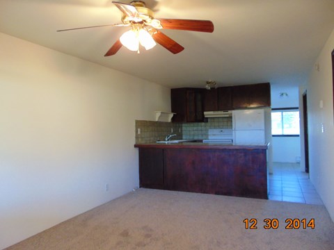 A room with a ceiling fan and a kitchen area in the background.