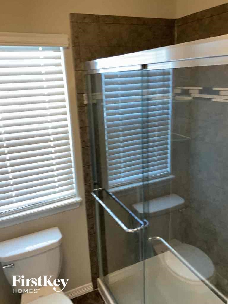 A bathroom with a toilet, a shower with a glass door and a window with blinds.