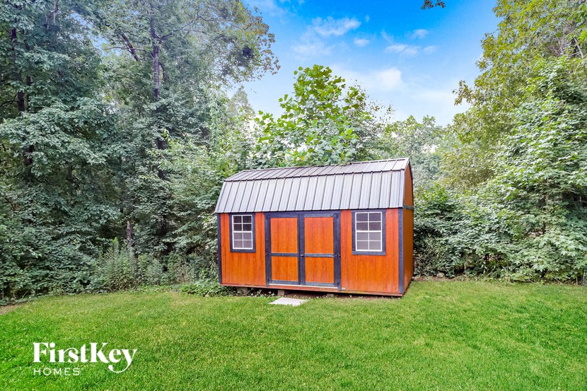 A small wooden shed with a metal roof is situated in a grassy area with trees in the background.