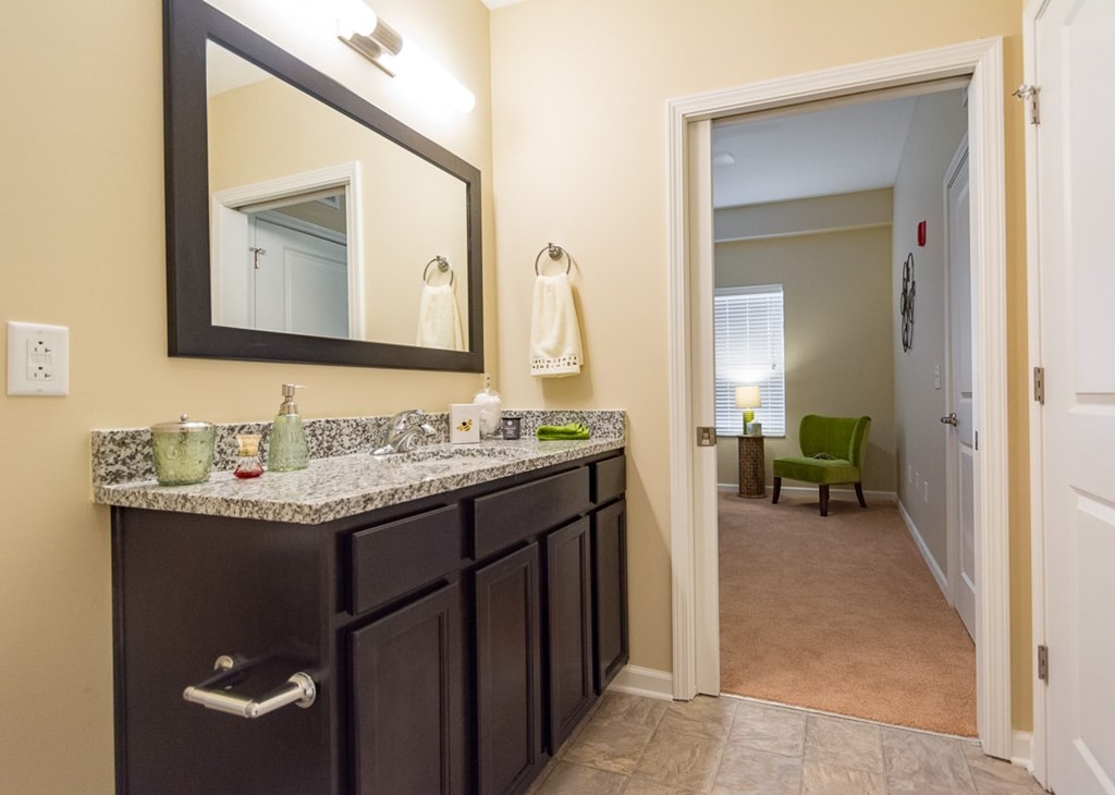 A bathroom with a large mirror above a vanity with a green chair in the hallway.