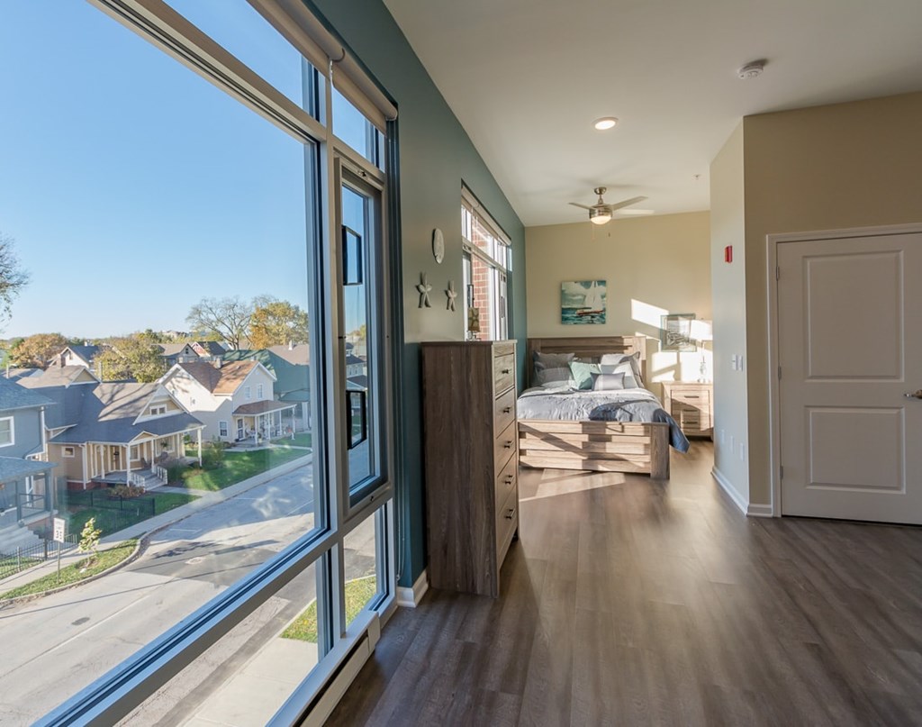 A bedroom with a large window overlooking a street with houses.