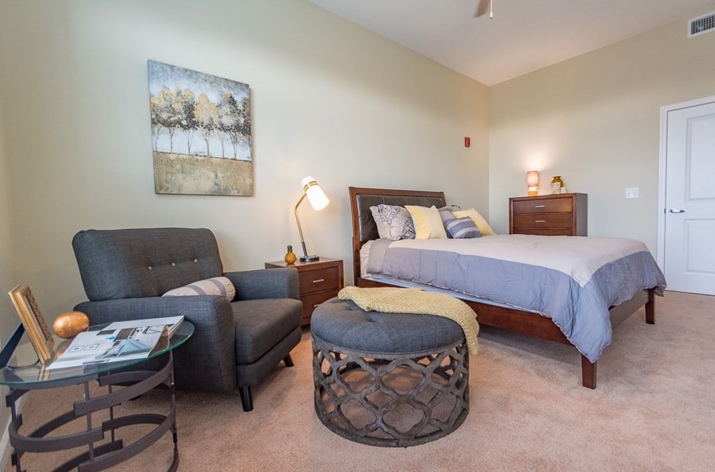 A bedroom with a bed, chair, ottoman, and painting.