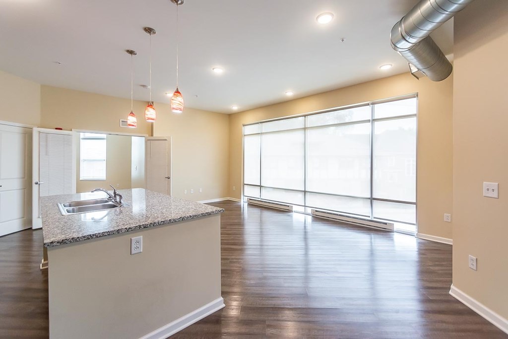 A kitchen island with a sink is in the middle of a large, empty room with wood floors and large windows.