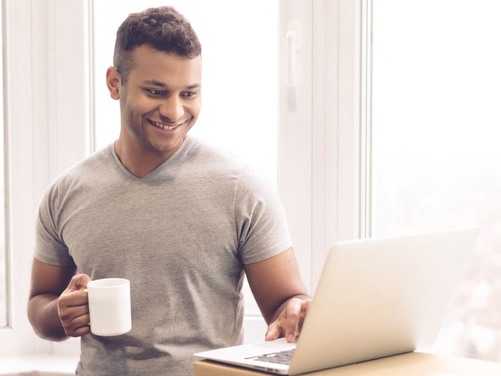 a man sitting at a table using a laptop computer and holding a mug