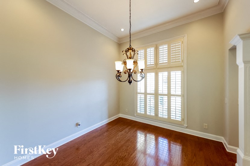 A chandelier hangs over a wooden floor in a room with white walls and a window with shutters.
