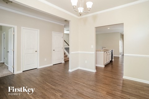 A spacious room with wooden floors and white walls, featuring a chandelier and a kitchen area in the distance.