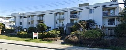 A white apartment building with balconies and bushes in front.