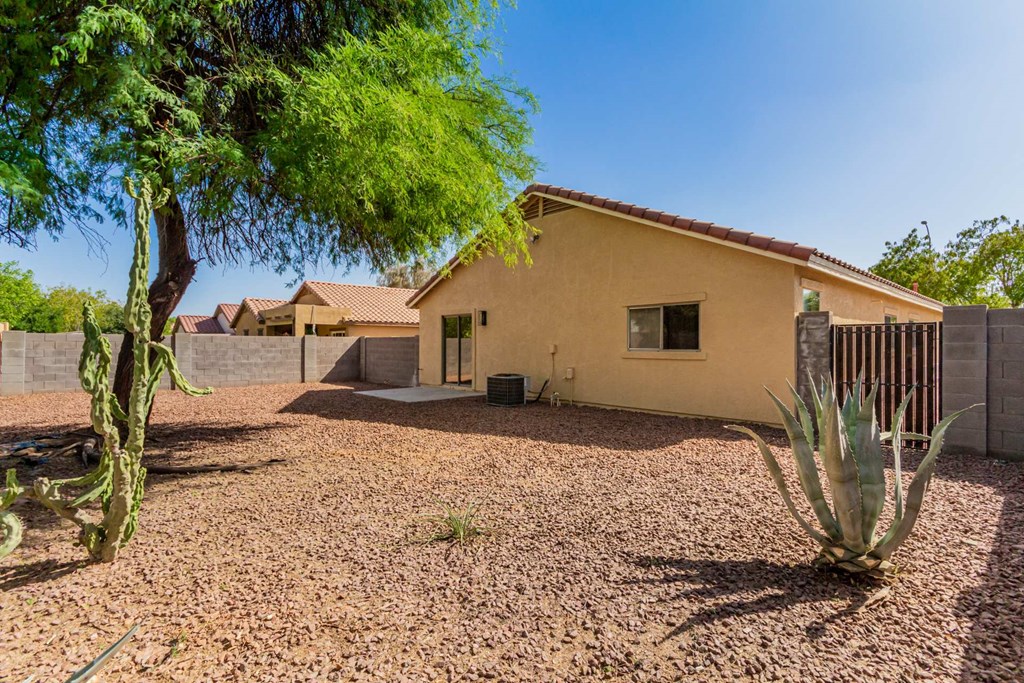 A house with a brown roof and a brown fence is surrounded by a gravel area with a cactus and a tree.