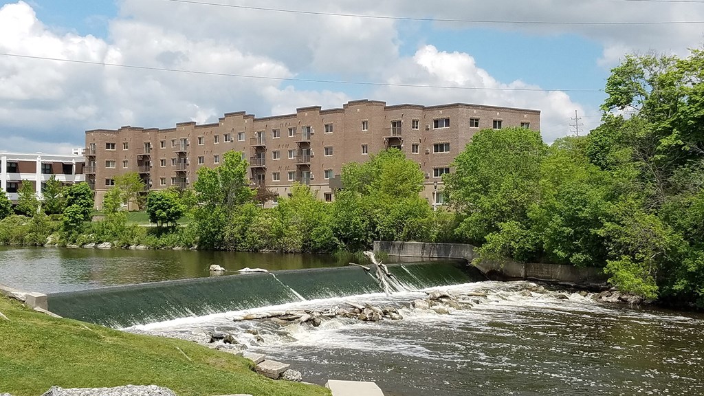 A large building sits next to a river with water flowing over a weir.